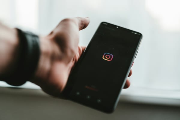 The Instagram logo on a black smartphone screen. The phone is in the hand of someone wearing a black bracelet