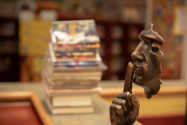 A photo of a sculpture of a man's face, in profile, holding a finger to his lips. In the background, books are out of focus