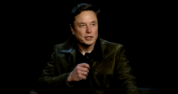 Musk wearing a black jacket, holding a microphone against a black backdrop