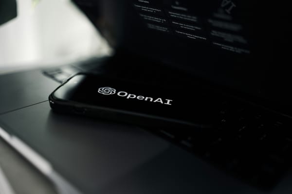 The OpenAI logo and wordmark on a black background on a smartphone laying on a laptop