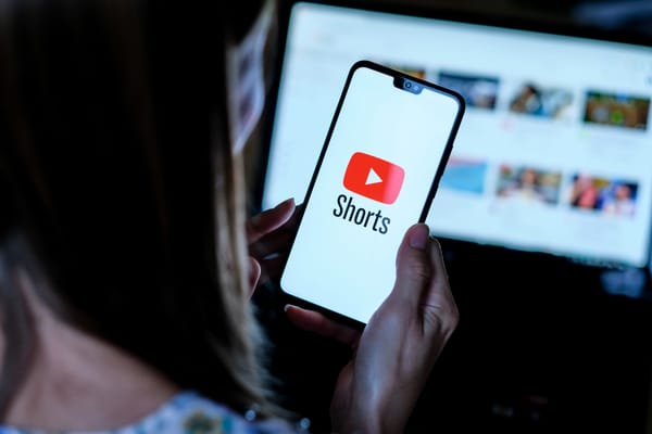 YouTube Shorts open on a phone held in a woman's hands. YouTube is open on a monitor in the background 