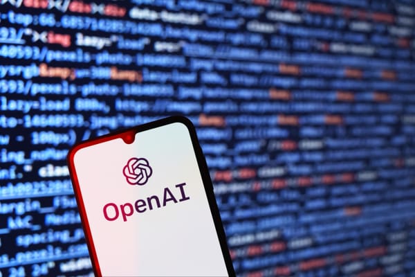 The OpenAI wordmark and logo shown on a smartphone with a white background against the backdrop of a monitor showing blurry red, white and blue code